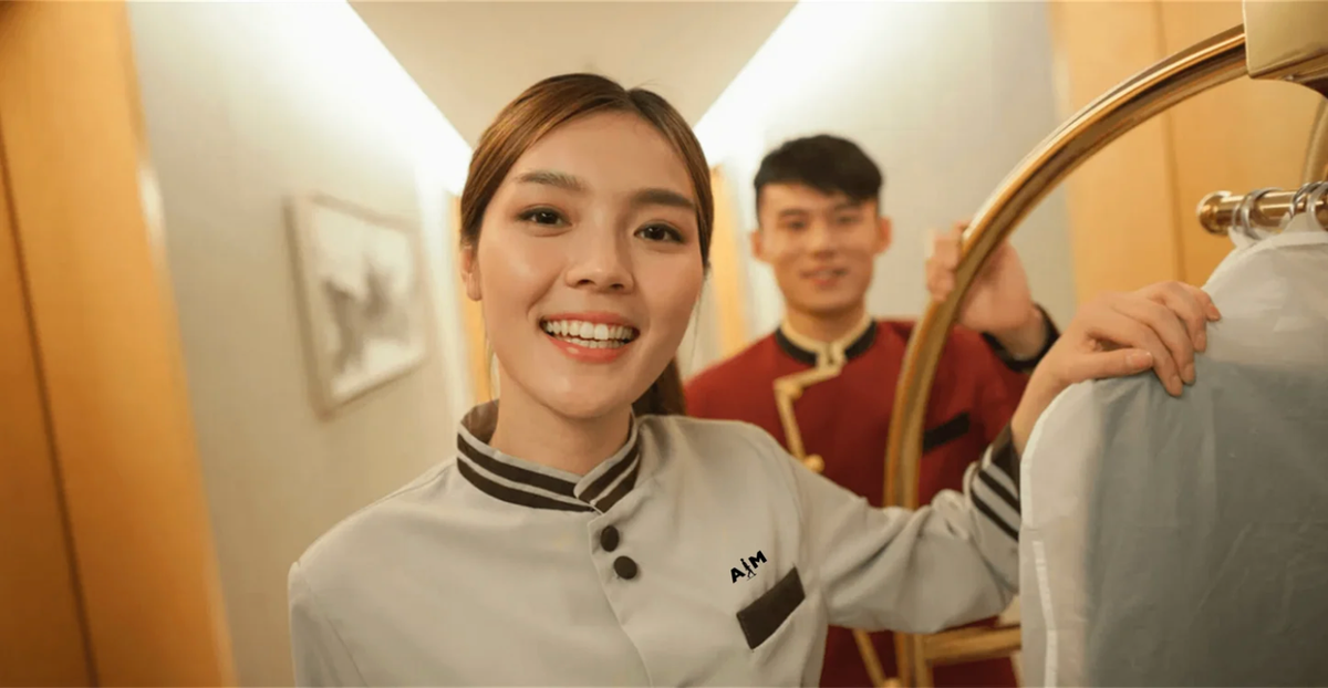 Two hotel colleagues in professional uniforms accompanying a garment trolley, with a small AIM logo on the light uniform. Illustrative hospitality concept.