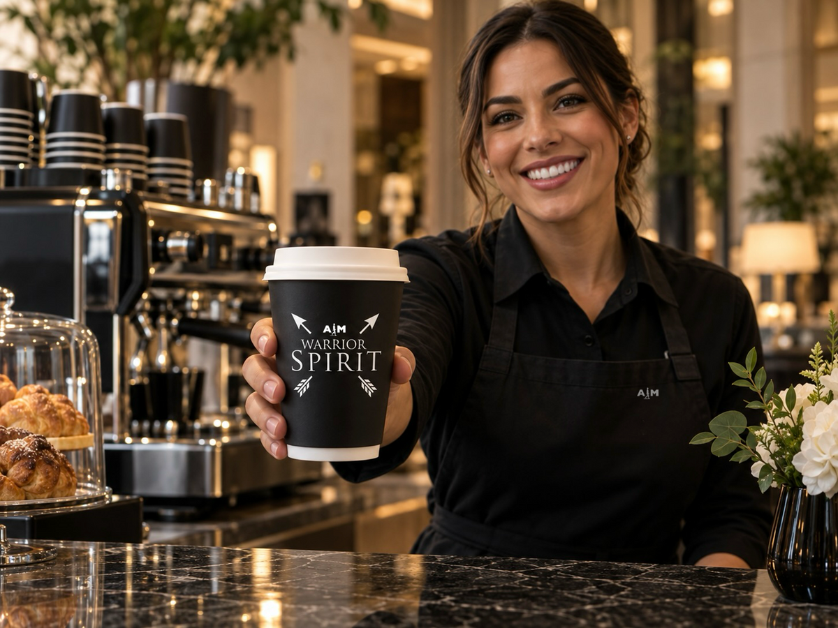 Illustrative AIM hospitality concept: a barista in a subtly branded apron handing a guest a latte in an AIM coffee cup.