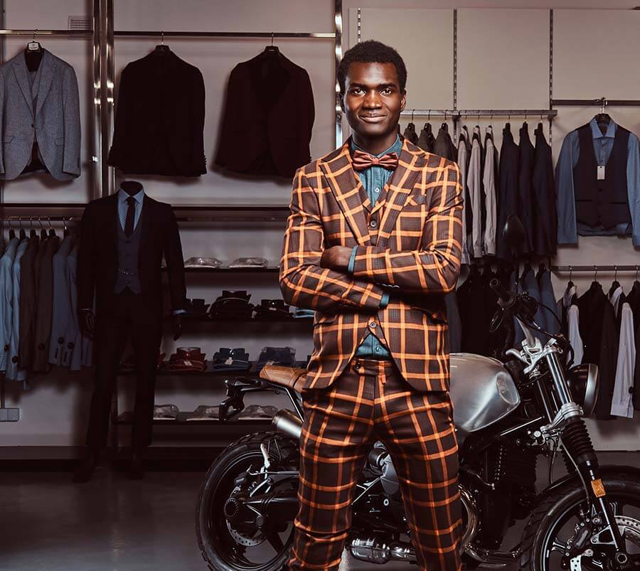 Confident African American man in a chic, patterned suit, standing in a boutique with a classic motorcycle, showcasing his unique style and self-esteem.