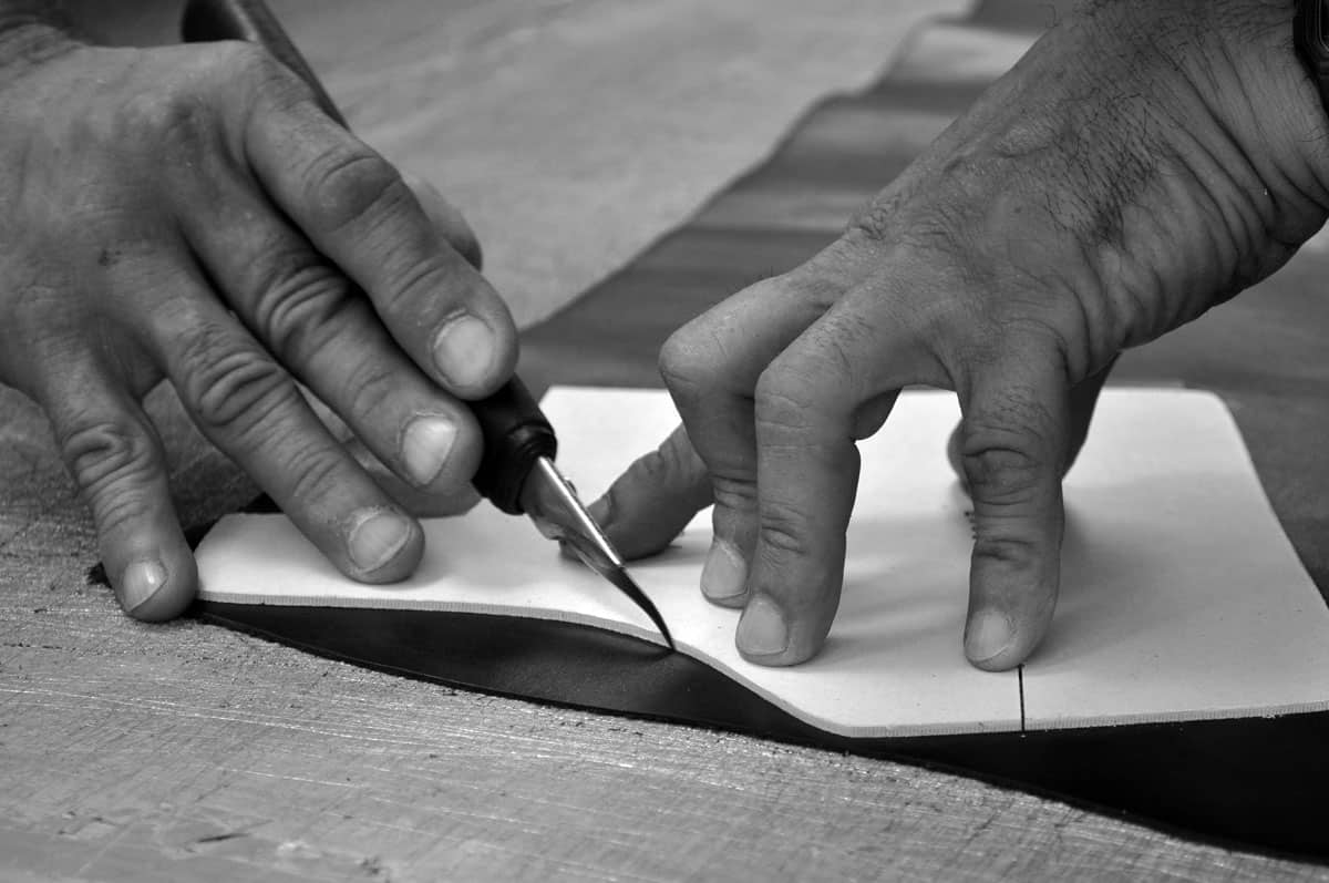 A black and white image capturing the hands of a skilled artisan meticulously cutting through material with a precision tool, highlighting the detailed and hands-on process of creating bespoke items.