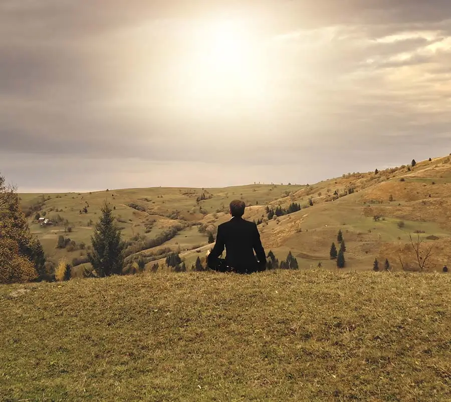 An individual sitting alone at the top of a grassy hill, overlooking a valley, under a bright sky, depicting a moment of reflection or solitude that can be important for one's mental health and emotional wellness.