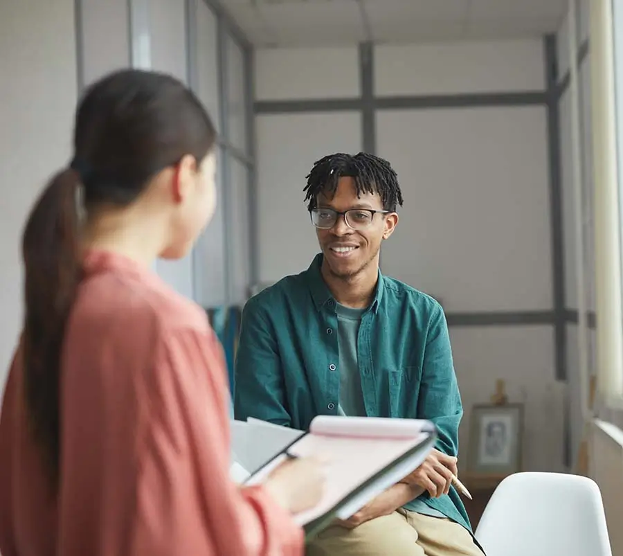 A man with glasses and dreadlocks, wearing a green shirt, smiling and having a conversation with a female colleague who is holding a tablet, in a setting that represents a supportive work environment contributing to mental health and emotional wellness.
