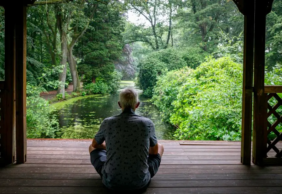 An adult man sits in contemplation under a wooden pergola, overlooking a tranquil garden pond, embodying the practice of mindfulness and presence in personal growth.