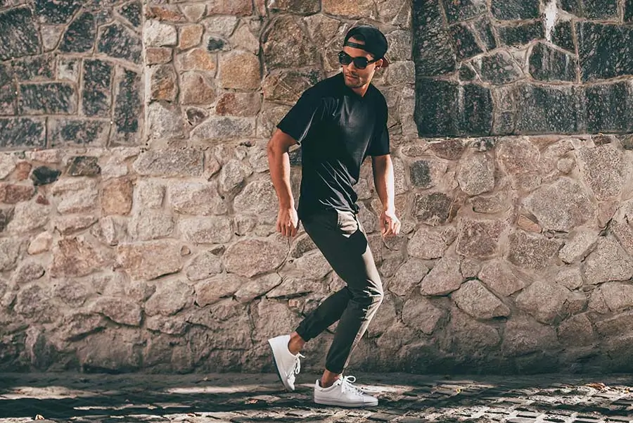 A confident young man in casual attire, including a black t-shirt, sunglasses, and white sneakers, walking along a historic cobblestone street, his poised demeanor reflecting a casual yet self-assured connection between sneakers and self-esteem.





