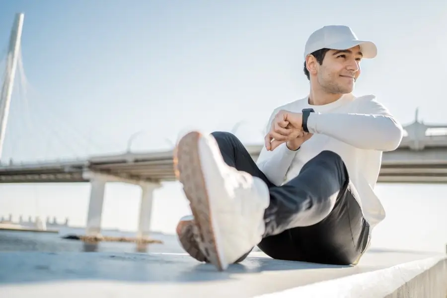 A relaxed man checking his fitness tracker by the water, showcasing the integration of technology and wellness in fostering a positive and healthy lifestyle.